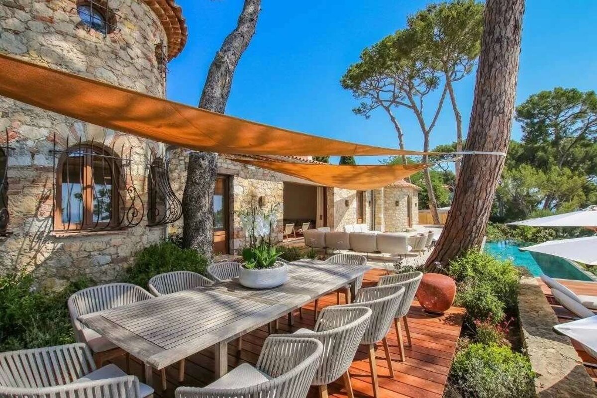 A luxurious stone villa patio features outdoor dining under sun shades, alongside a swimming pool and lush trees on a sunny day.