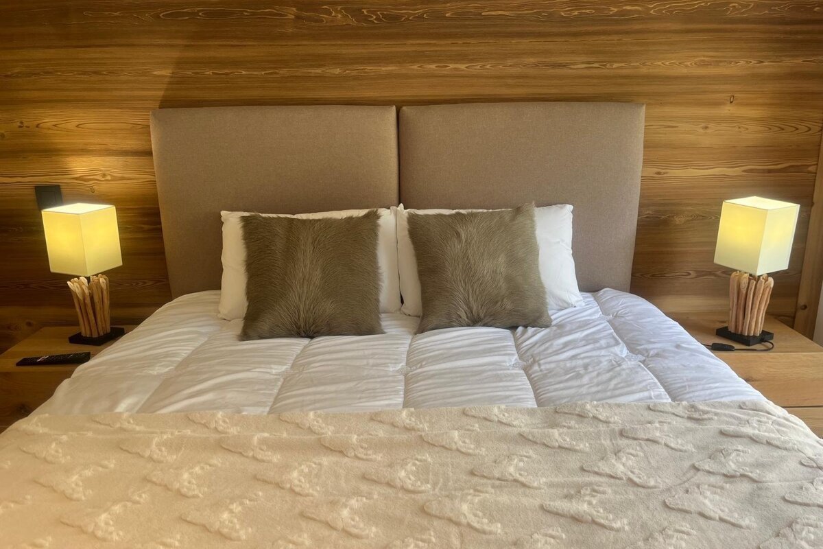 Cozy bed with white linens and brown fur pillows, flanked by matching stick-base lamps against a warm wood-paneled wall.