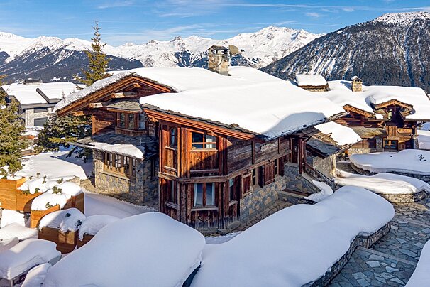 Stunning wooden and stone chalets covered in deep snow, with majestic snow-capped mountains and evergreen trees under a clear blue sky.