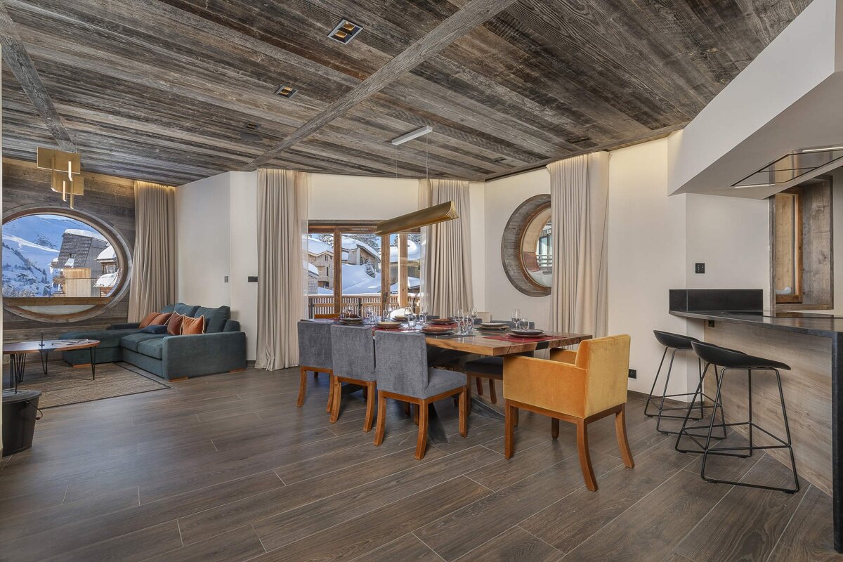 Modern ski chalet interior: rustic wood ceiling, large circular window with mountain view, open-plan living/dining, and sleek kitchen bar.