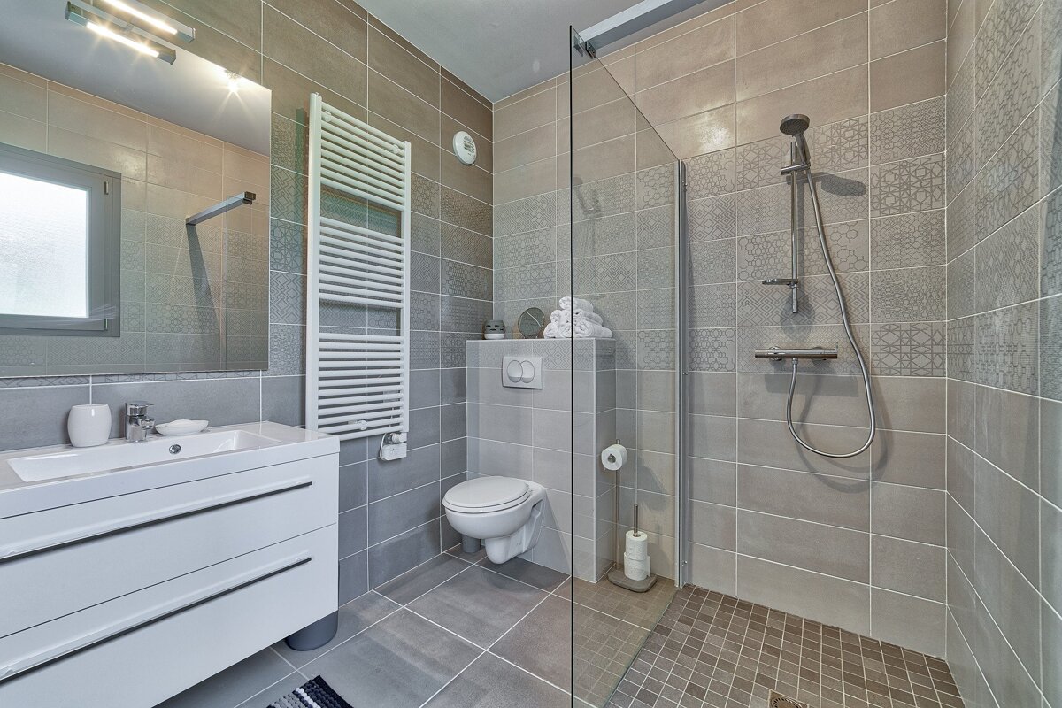 A modern, grey-tiled bathroom with a walk-in shower, floating toilet, white vanity, and towel warmer, featuring patterned accent tiles.