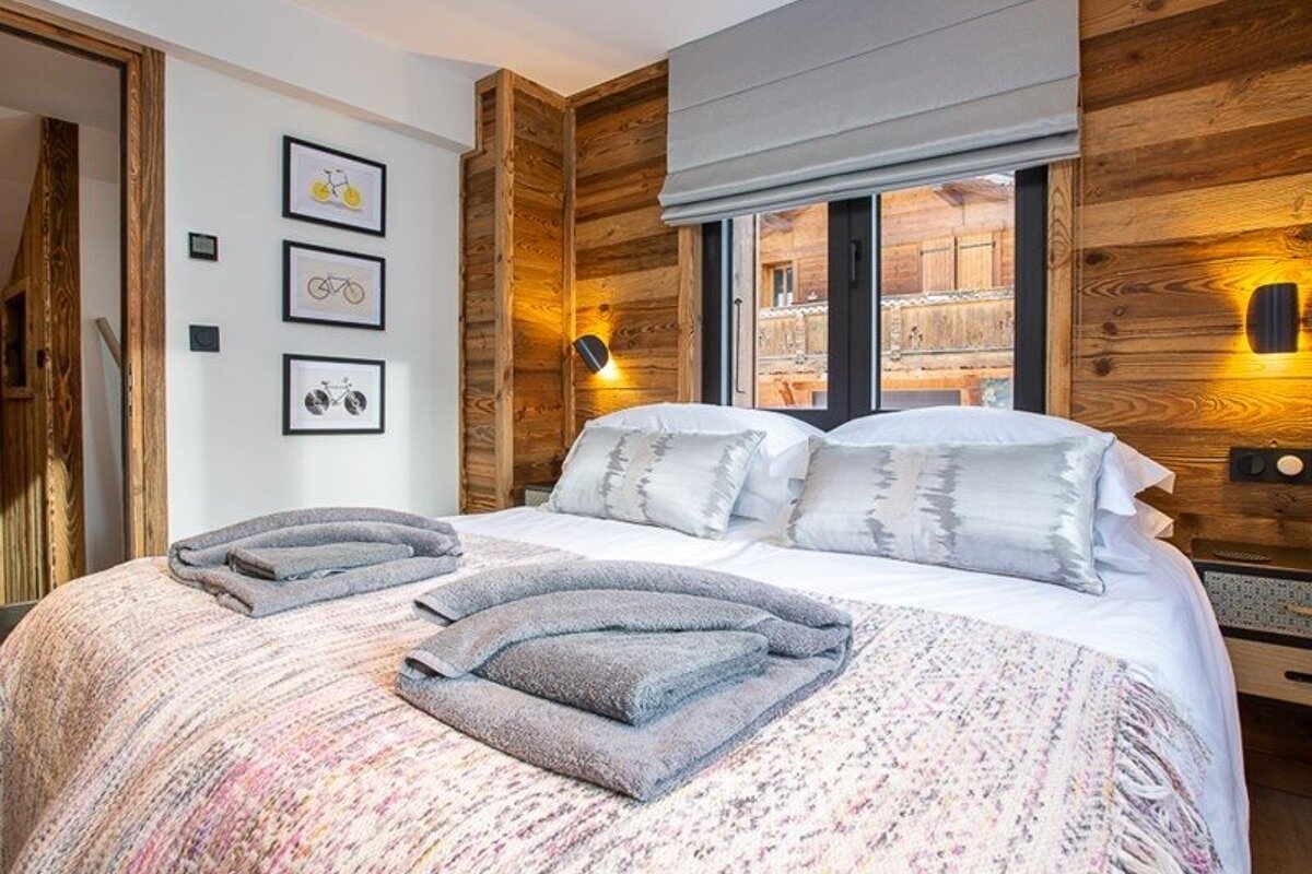 Cozy chalet bedroom with wood-paneled walls, a neat double bed with folded grey towels, framed bicycle art, and a small window.