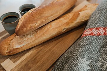 Two golden baguettes and two cups of coffee rest on a wooden board, next to a grey blanket with a red pattern.