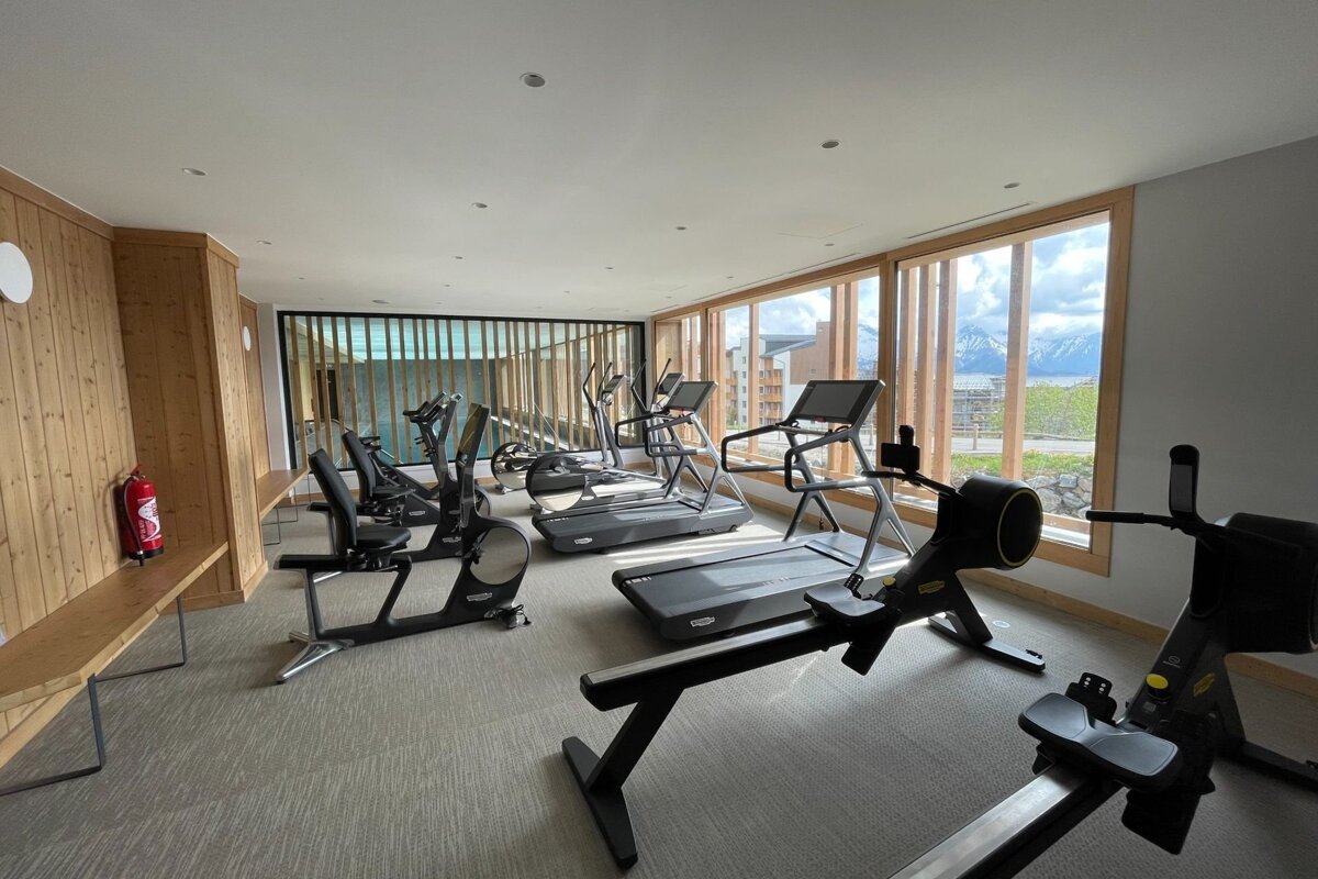 A gym equipped with treadmills, rowing machines, and stationary bikes, featuring large windows with scenic mountain views.