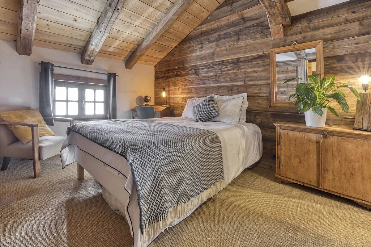 Cozy attic bedroom with rustic wooden walls & ceiling beams. Features a bed, armchair by a window, and wooden furniture with a plant.