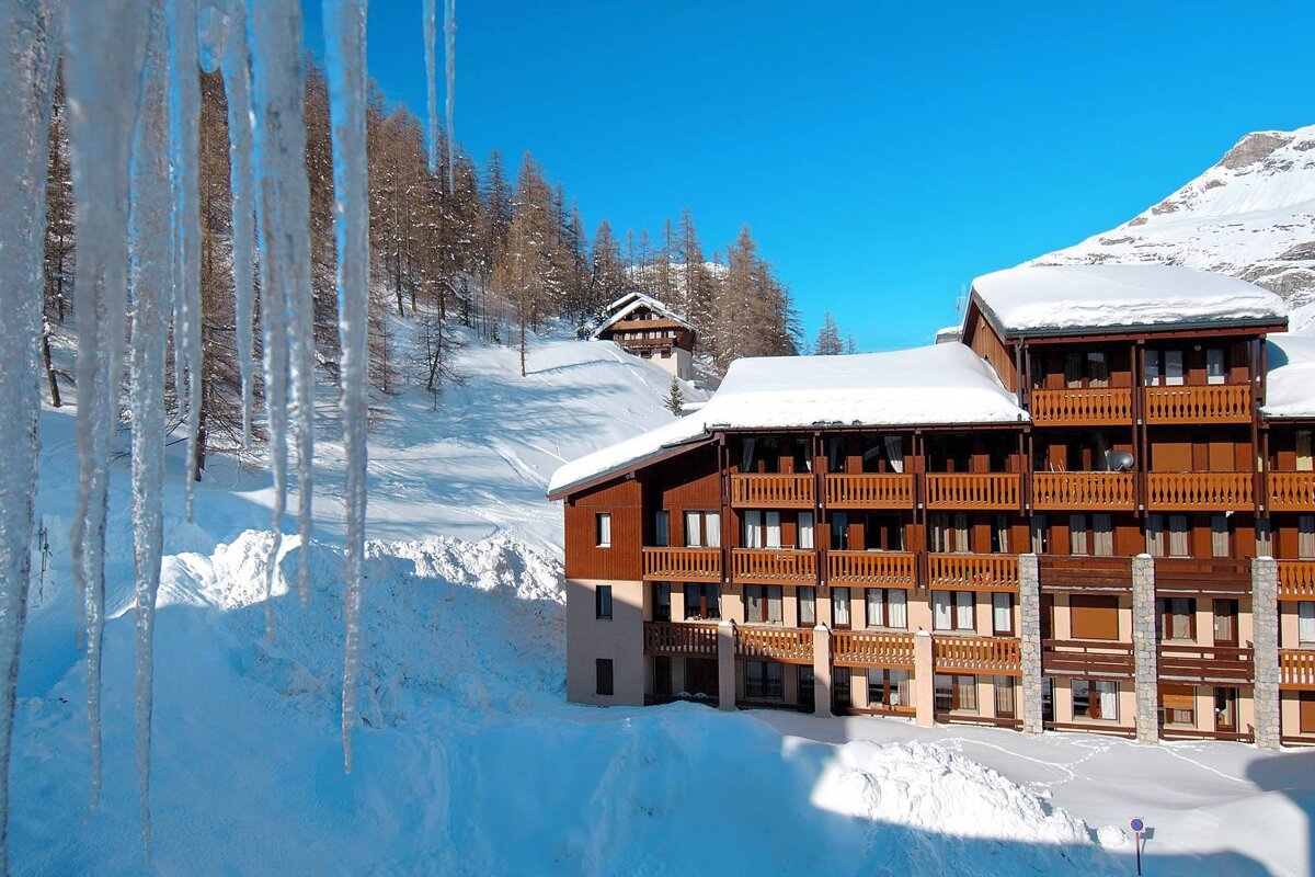 Long icicles frame a snowy mountain scene with a large wooden resort building, distant trees, and a bright blue sky.