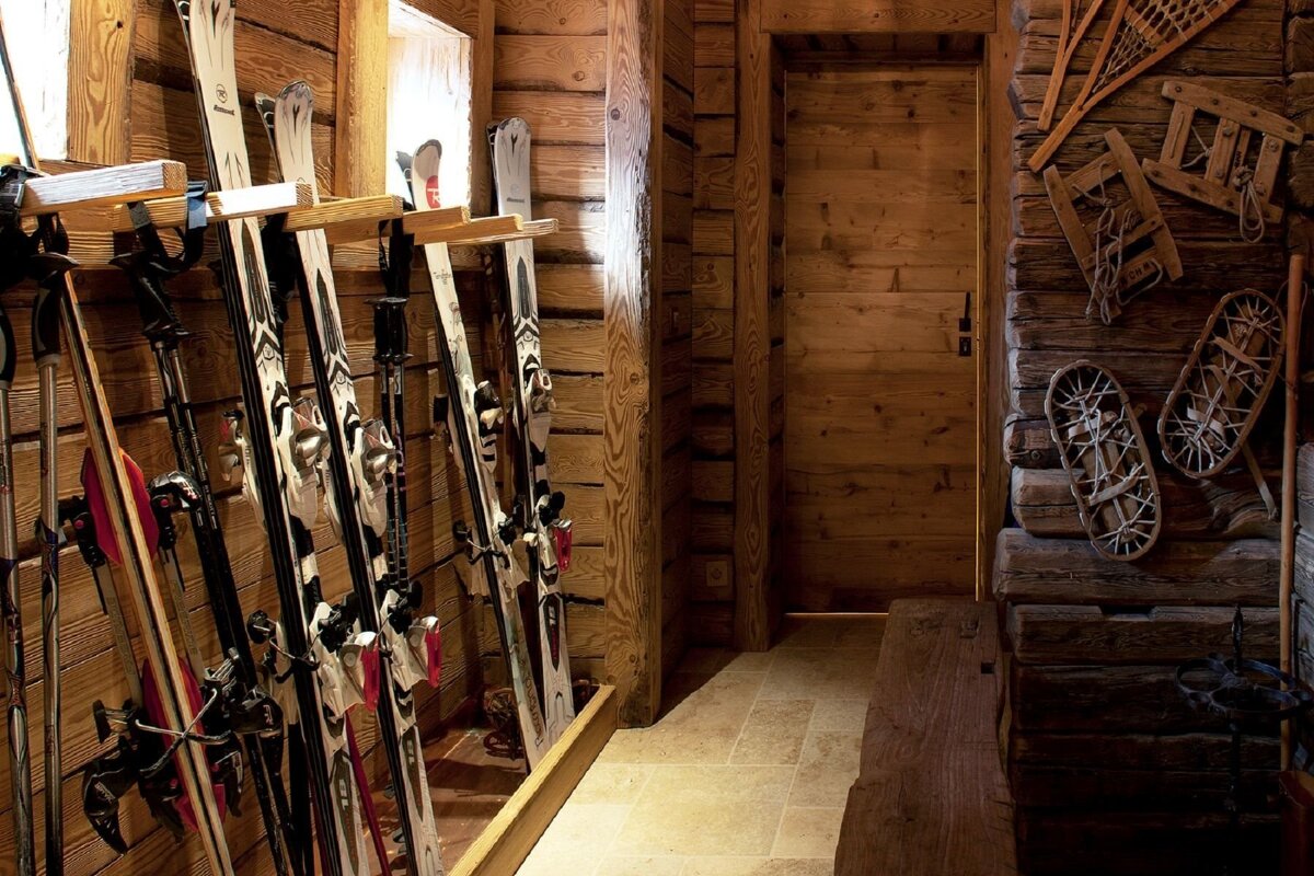 A rustic wooden ski room features multiple skis and poles neatly stored, with vintage snowshoes and sleds adorning a timber wall. A wooden door and bench complete the cozy scene.