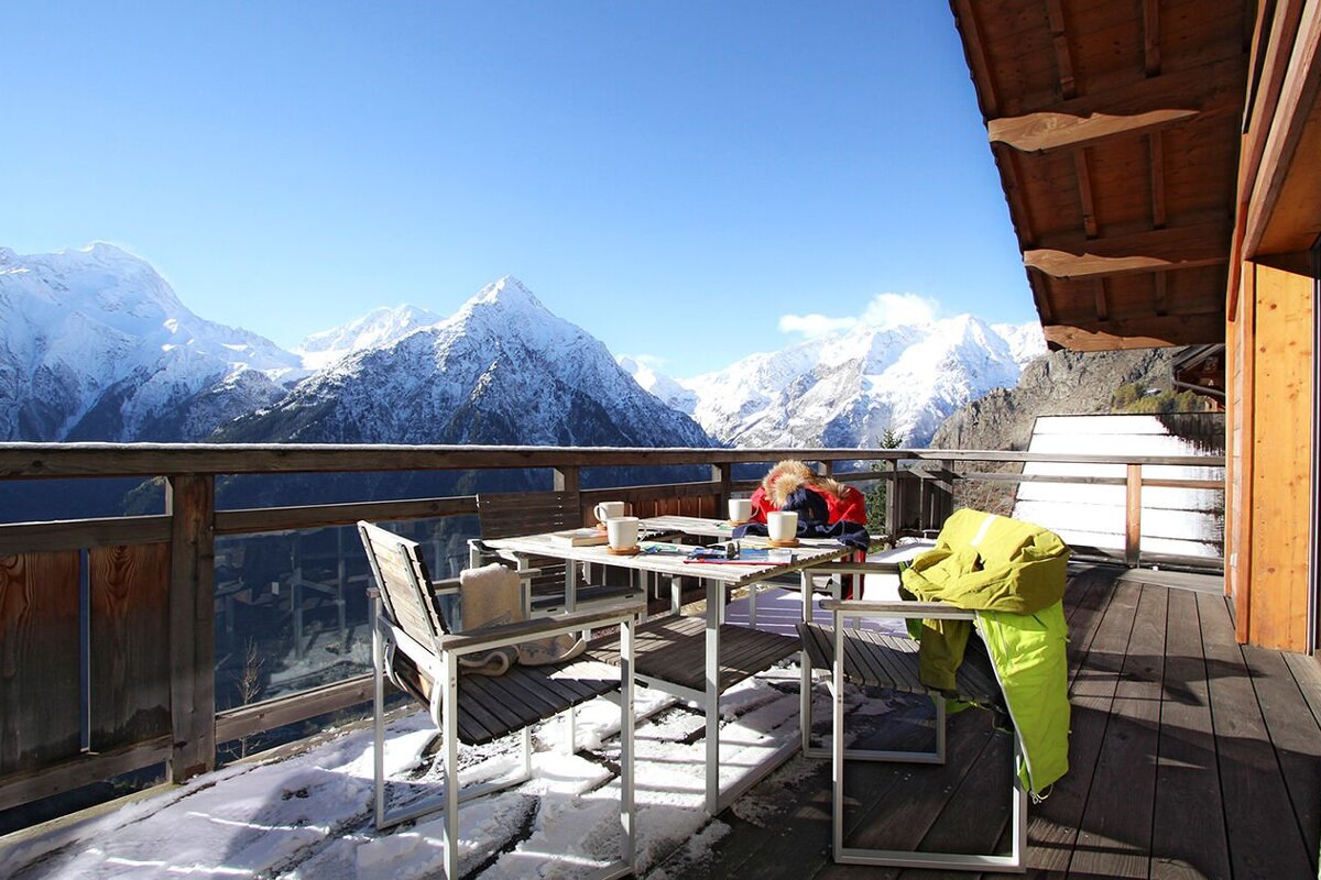 A woman sits at a table on a balcony overlooking snowy mountains