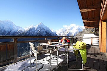 A woman sits at a table on a balcony overlooking snowy mountains