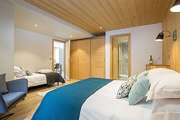 Modern bedroom with wood ceilings, a large bed with a blue throw, a single bed, and an open doorway to a bathroom.