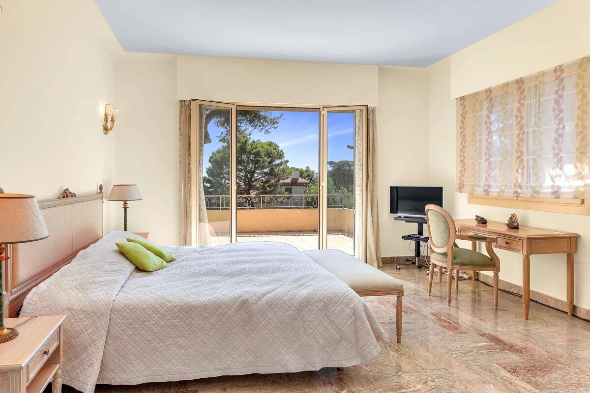 A spacious, light-filled bedroom featuring a large bed, private balcony access, a desk, TV, and patterned marble floors.
