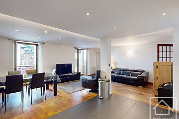 A modern open-plan living and dining room with wooden floors, black leather seating, a TV setup, and recessed ceiling lighting.