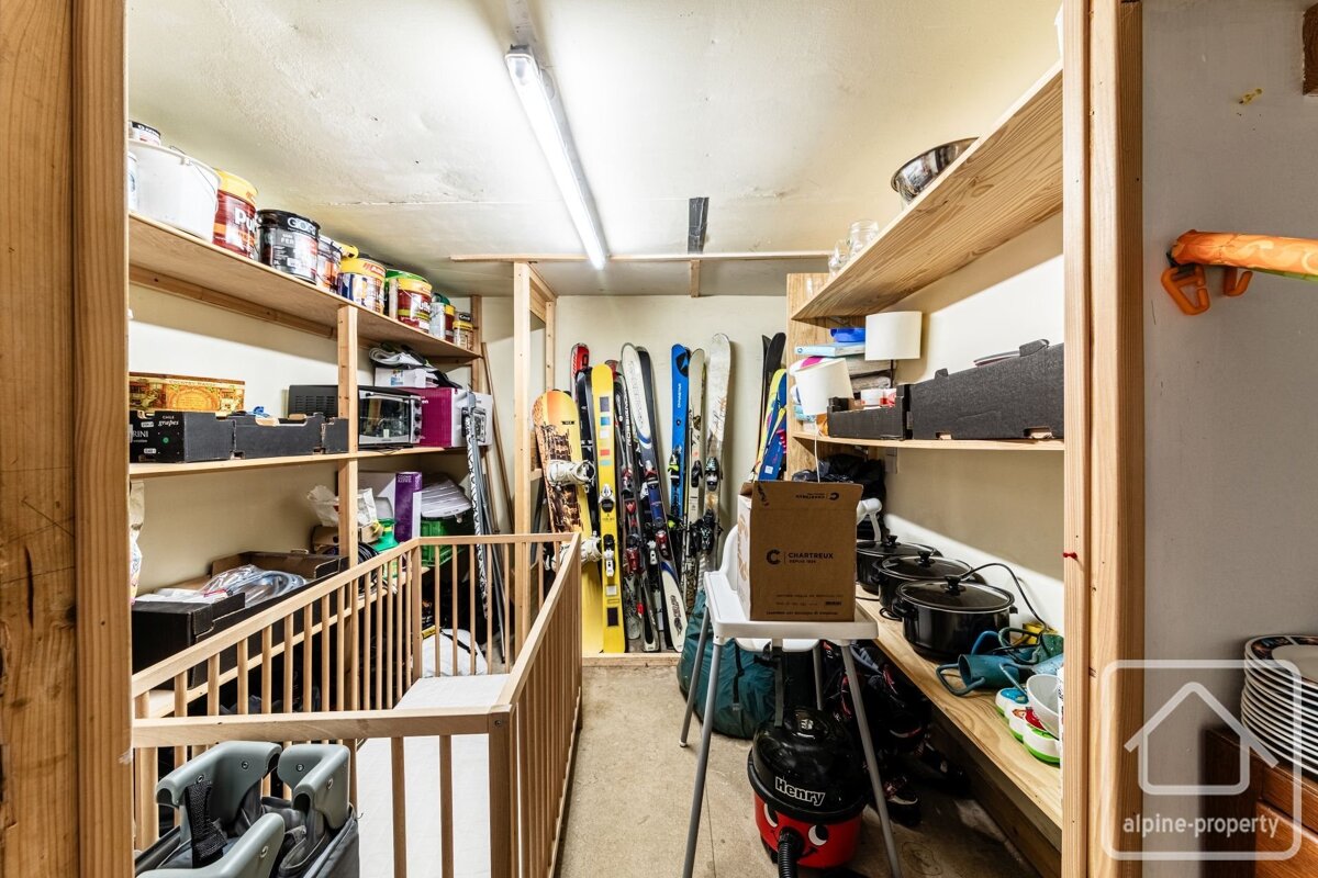 A cluttered storage room with wooden shelves packed with paint, kitchenware, boxes, and a Henry vacuum cleaner. Skis and snowboards stand along one wall, with a baby gate visible.