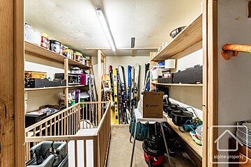 A cluttered storage room with wooden shelves packed with paint, kitchenware, boxes, and a Henry vacuum cleaner. Skis and snowboards stand along one wall, with a baby gate visible.
