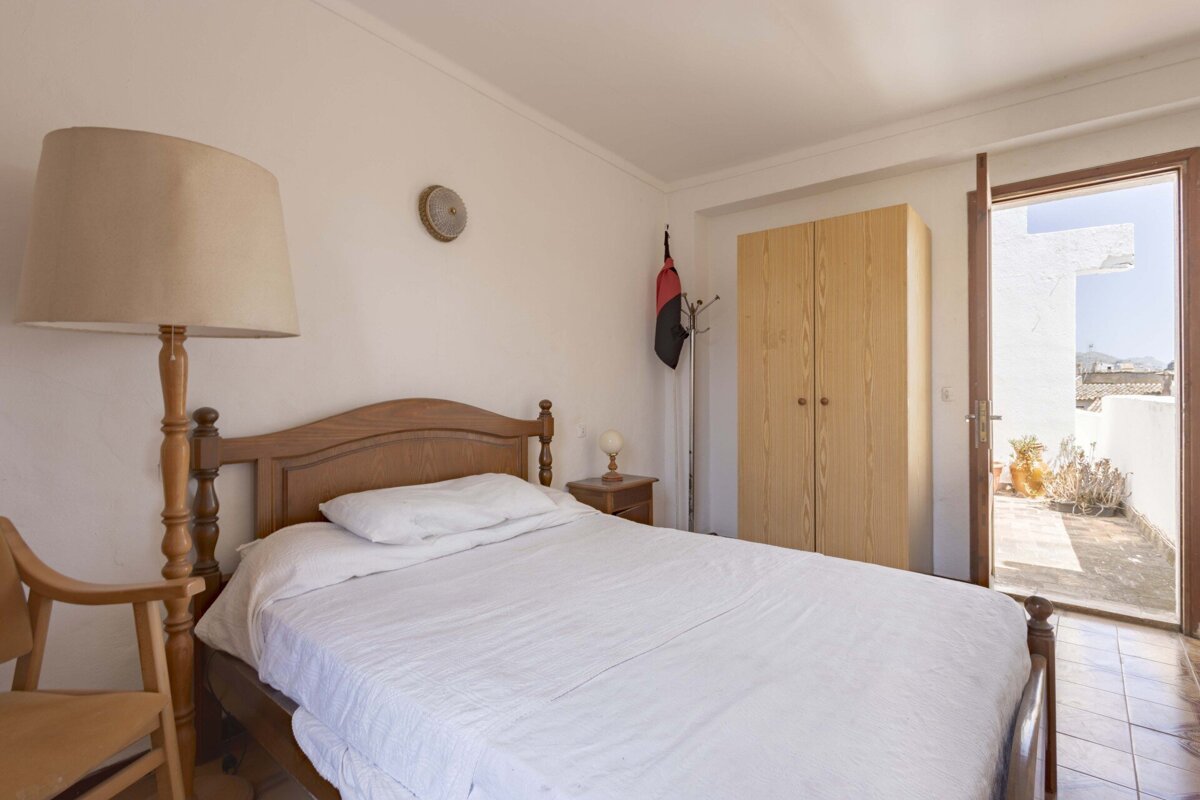 A bright bedroom with a wooden bed, white bedding, floor lamp, and a wardrobe. A door opens to a tiled outdoor terrace.