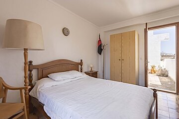A bright bedroom with a wooden bed, white bedding, floor lamp, and a wardrobe. A door opens to a tiled outdoor terrace.