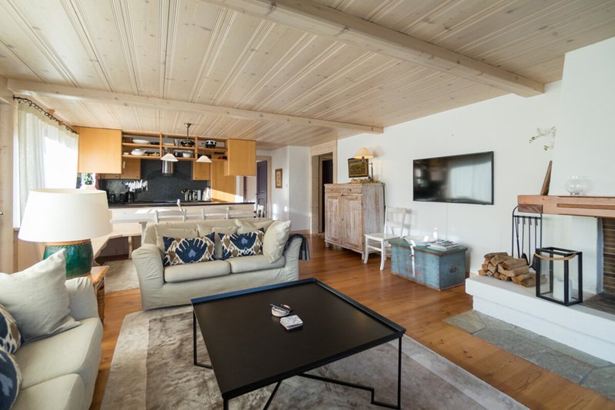 A cozy, rustic living room featuring a wood-paneled ceiling, beige sofas, a black coffee table, a fireplace, and a kitchen in the background.