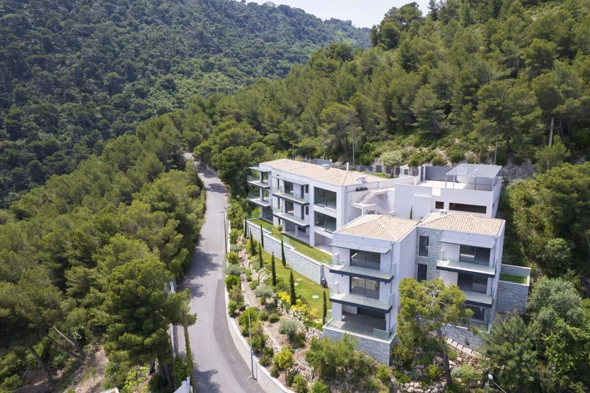Aerial view of modern white buildings with terraced gardens and terracotta roofs, nestled in a lush green mountainous forest alongside a winding road.