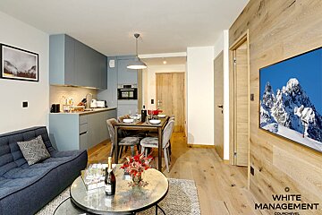 A stylish open-plan apartment featuring a sleek blue kitchen, dining table, cozy sofa, and a large TV showing a snowy mountain landscape on a wood-paneled wall.