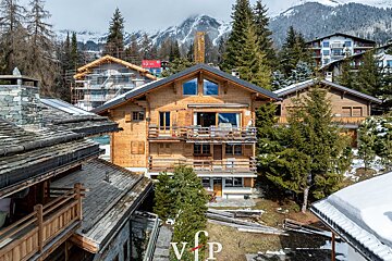 Cozy wooden alpine chalets surrounded by pine trees, set against snow-covered mountains.