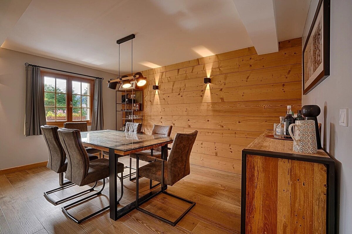 A rustic-modern dining room with wooden walls, an industrial-style table and chairs, and a window view. Warm lighting creates a cozy ambiance.