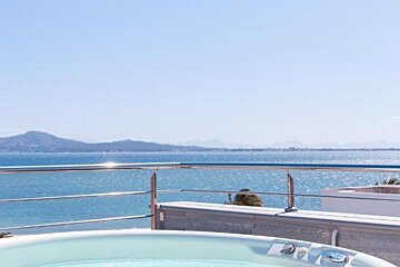 A hot tub on a balcony overlooks a sparkling blue sea and distant mountains under a clear sky.