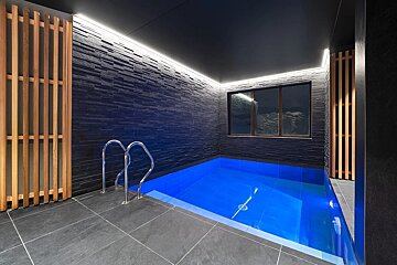 A modern indoor pool with bright blue water, dark slate walls, wooden accents, strip lighting, and a window looking out to a dark scene.