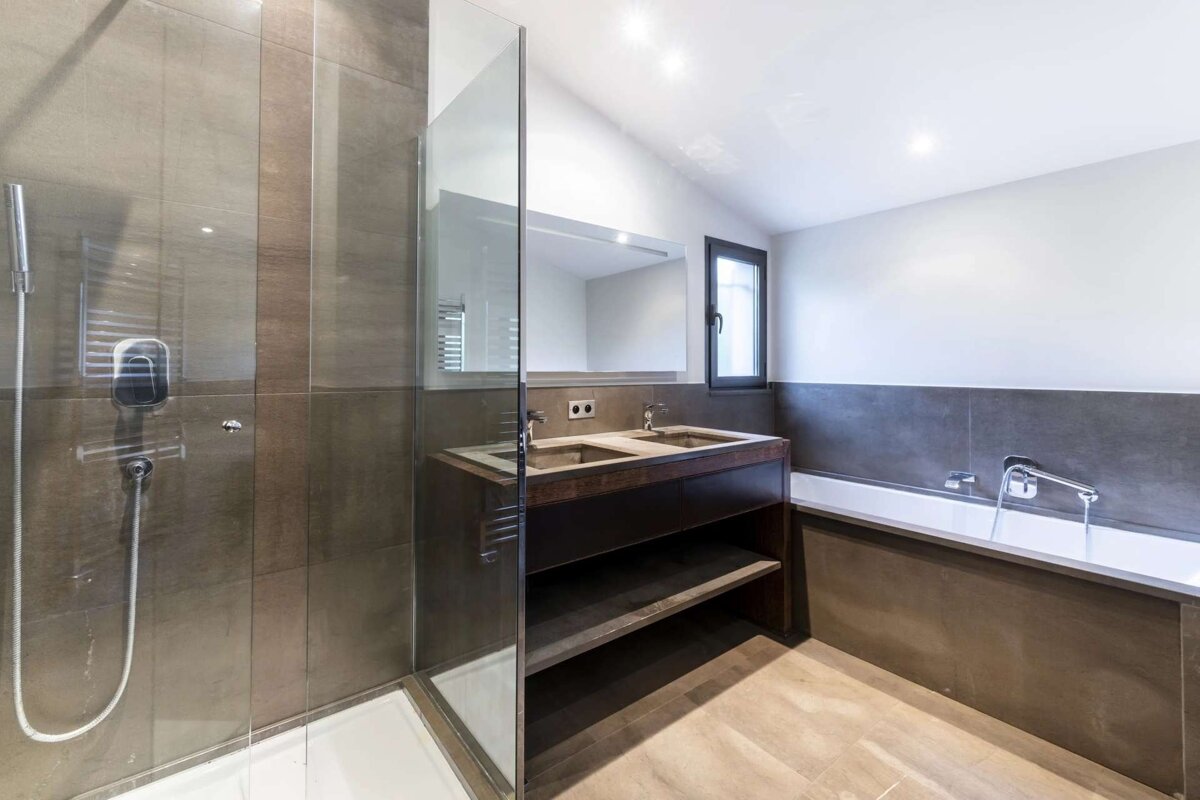 A modern bathroom featuring a glass shower, double vanity, and built-in bathtub. It has neutral grey tiles, white walls, and a window.