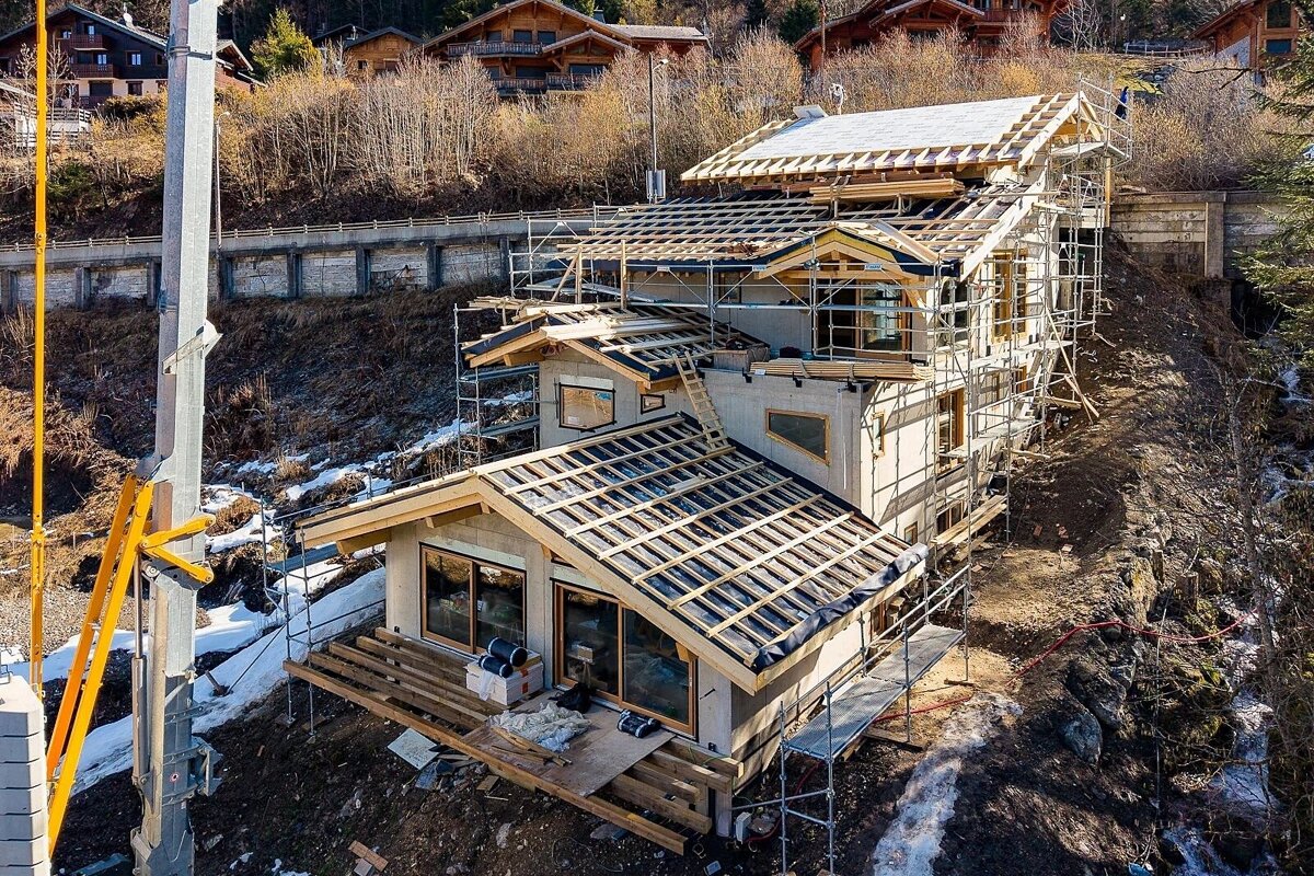 A large multi-level house is under construction on a steep, snow-dusted hillside, surrounded by scaffolding and framed roofs. Other chalets dot the background.