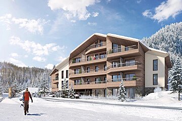 A modern wooden ski resort building is nestled in a snowy mountain landscape. A person carrying a snowboard walks on a snow-covered path.