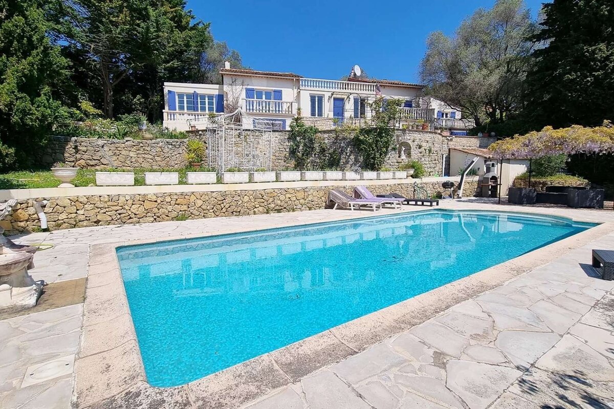 A beautiful large house with blue shutters and stone terracing overlooks a sparkling blue swimming pool, surrounded by lush greenery under a clear sky.
