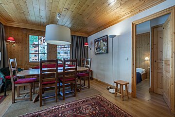 Cozy, wood-paneled dining room with a large table, red-cushioned chairs, window view, and an open doorway leading to a bedroom.