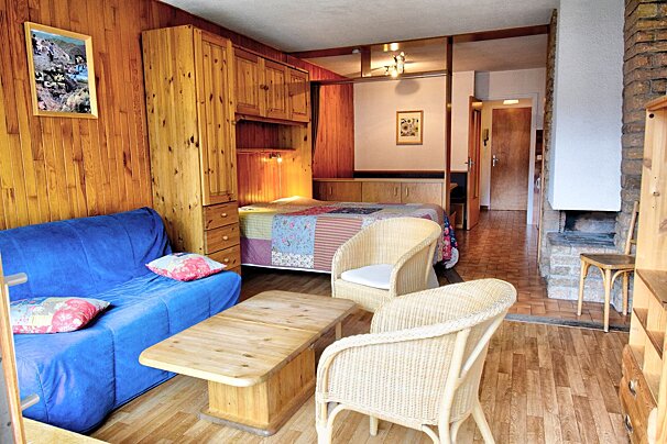 A cozy wood-paneled living area with a blue sofa, wicker armchairs, a wooden coffee table, and a bed with a quilt in the background.