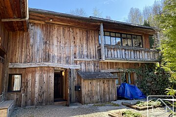 Weathered wooden building with an open door, upper balcony, large windows, and a small shed. Trees and blue tarp.