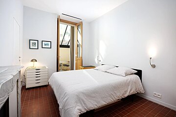 A clean, bright bedroom features white walls, terracotta tiled floor, a bed with white bedding, and an open door leading to an outdoor patio.