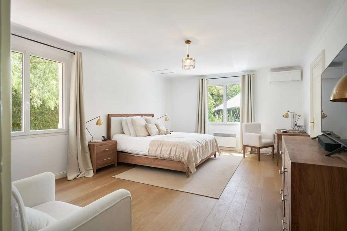 Bright, modern bedroom with white walls, wood floor, large bed, two windows overlooking greenery, and minimalist decor.