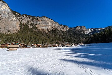 A snow-covered village sits at the base of forested mountains under a bright blue sky, with ski tracks visible in the foreground.