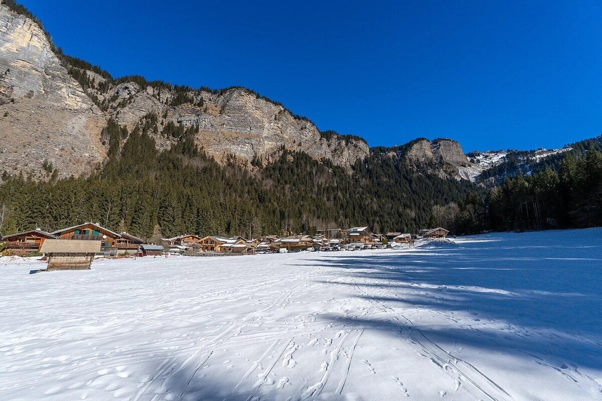 Snow-covered field with an alpine village of chalets beneath steep, forested rocky mountains under a clear blue sky.