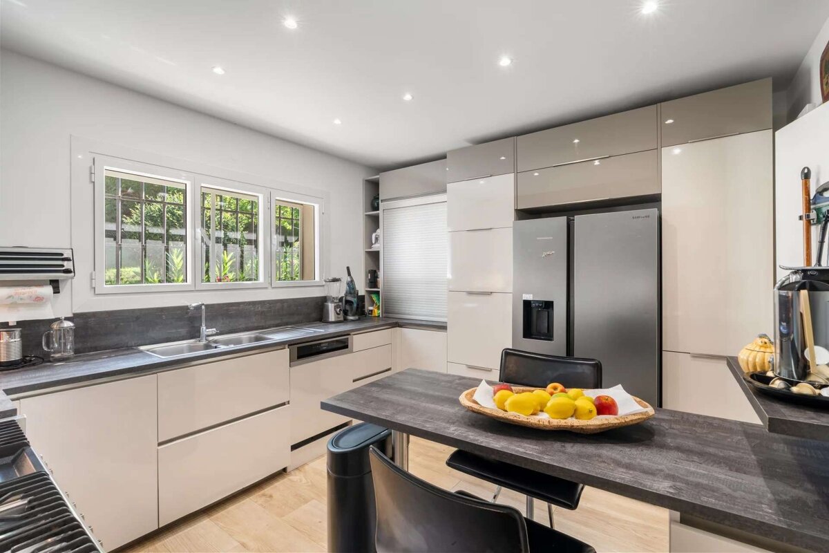 A modern kitchen with light and grey cabinets, stainless steel appliances, dark countertops, and a dining peninsula with a fruit bowl. A window overlooks greenery.