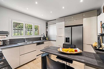 A modern kitchen with light and grey cabinets, stainless steel appliances, dark countertops, and a dining peninsula with a fruit bowl. A window overlooks greenery.