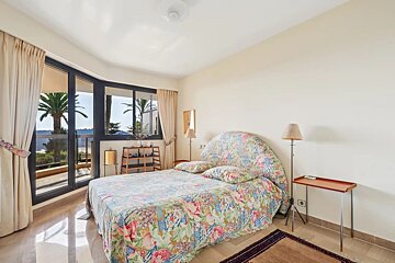 Bright bedroom with a floral bed, beige walls, and glass doors opening to a balcony with palm trees.