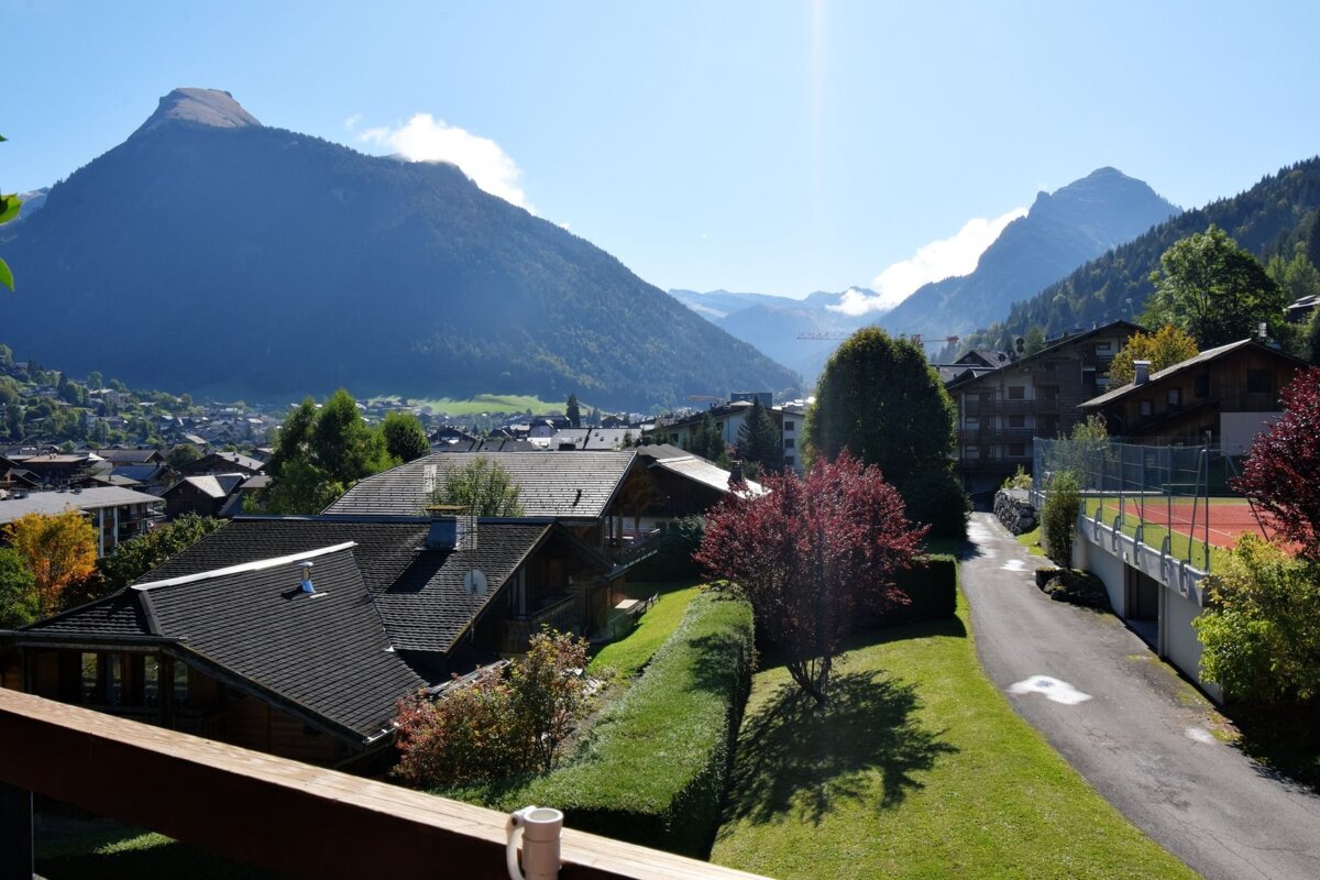 Panoramic view of a sunny mountain village in a valley, framed by towering peaks. Houses, lush greenery, a road, and a tennis court are visible.