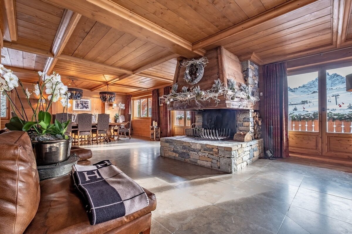 Rustic luxury ski chalet living room with wood paneling, a large stone fireplace, dining area, and a stunning snowy mountain view.