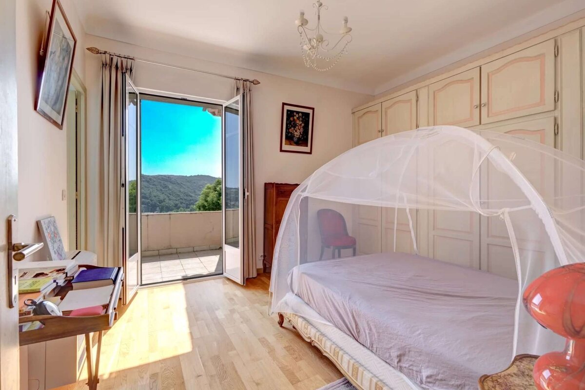 A bright bedroom with a bed under a mosquito net, hardwood floors, a built-in wardrobe, and French doors opening to a balcony with a panoramic mountain view.