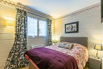 A rustic yet cozy bedroom featuring light wood paneling, a patterned bedspread with a purple throw, themed curtains, a window, and artwork.
