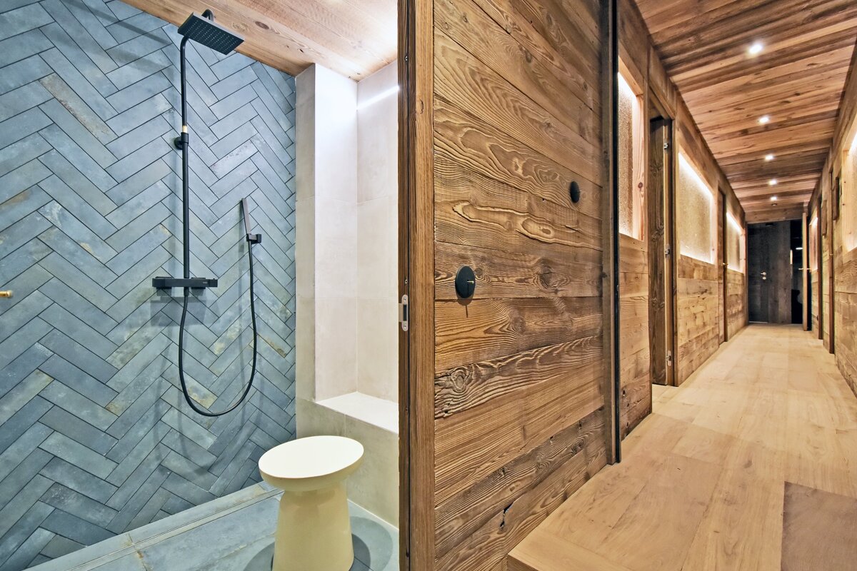 A stylish bathroom with blue herringbone tile shower and black fixtures, adjoins a rustic-chic wooden hallway with warm recessed lighting.