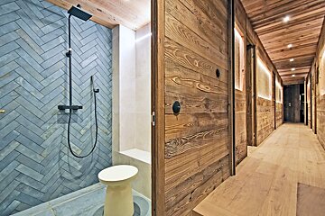 A stylish bathroom with blue herringbone tile shower and black fixtures, adjoins a rustic-chic wooden hallway with warm recessed lighting.
