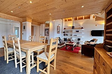 Cozy cabin interior with a wooden dining table and chairs in the foreground and a warm living area in the background.