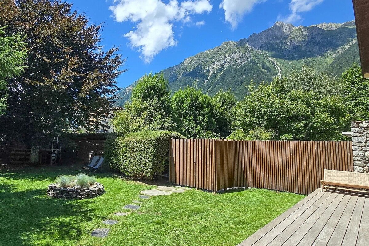 Sunny backyard with a green lawn and wooden deck, offering a scenic view of towering alpine mountains under a bright blue sky.