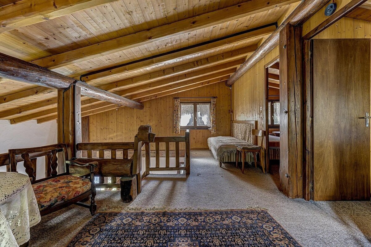 A rustic, wood-paneled attic bedroom with a sloped ceiling, exposed beams, a single bed by a window, and traditional wooden furniture.